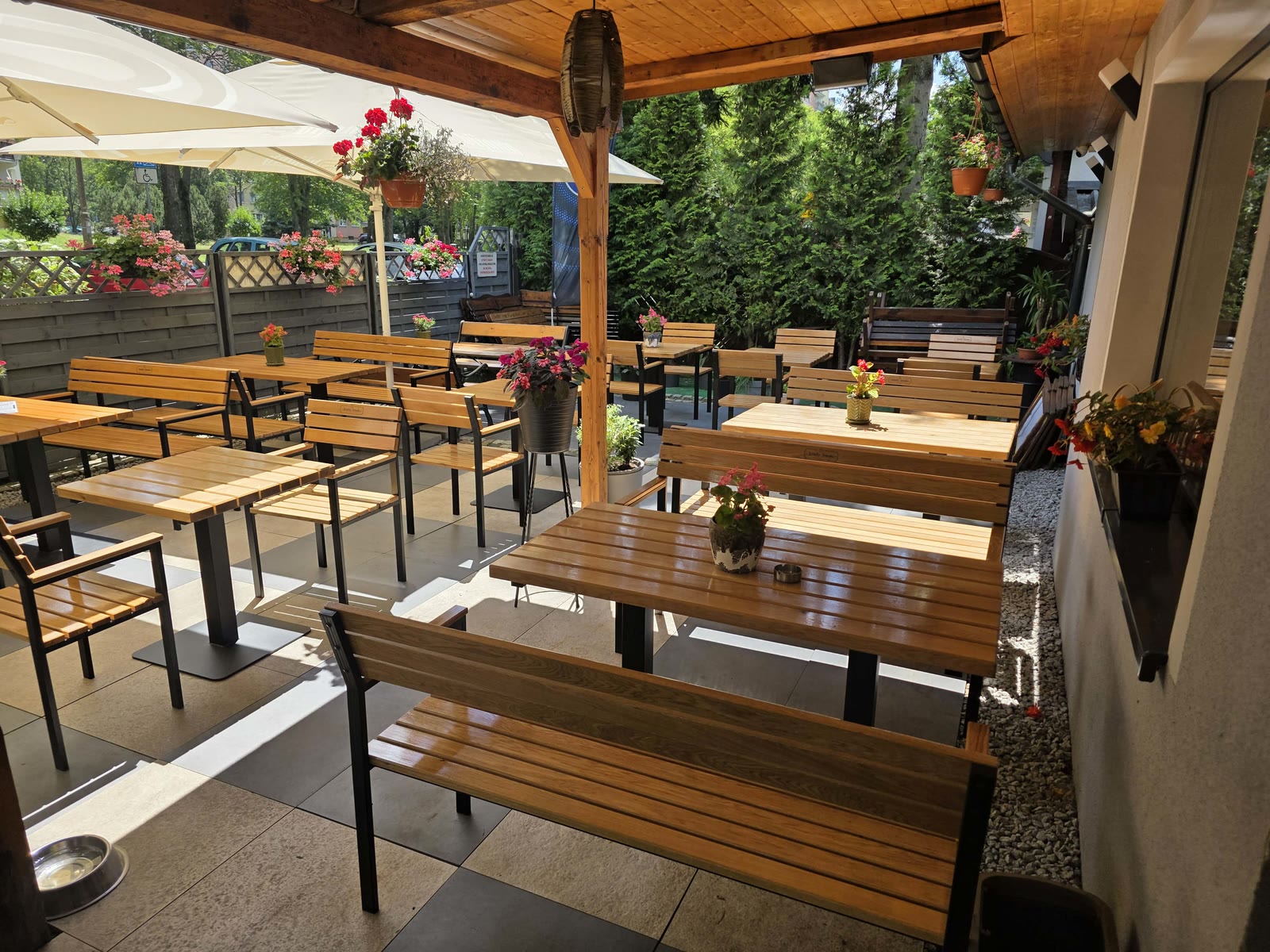 Letni ogródek restauracji z drewnianymi stolikami, kwiatami i zielenią.